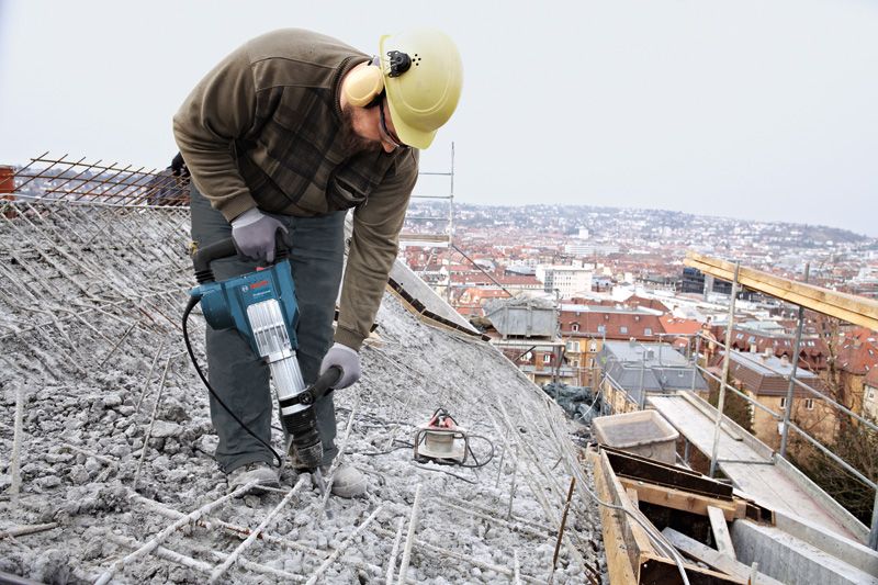 안전장비를 착용한 사람이 철거 망치를 사용해 옥상의 콘크리트를 깨고 있습니다.