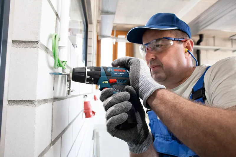 안전 장비를 착용한 사람이 벽에서 무선 콤비 드릴을 사용하고 있습니다.