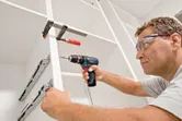 안전장비를 착용한 사람이 무선 드릴을 사용하여 캐비닛을 조립하고 있습니다.
