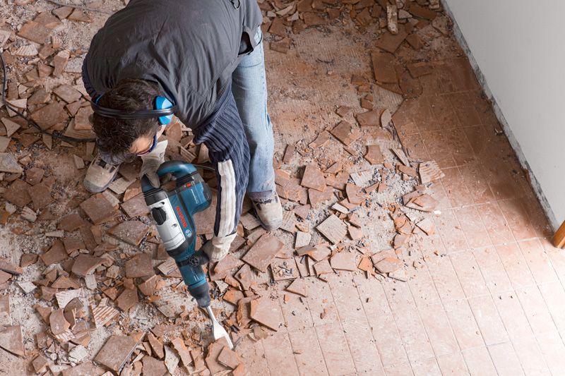 안전장비를 착용한 사람이 파괴망치를 이용해 바닥 타일을 부수고 있습니다.