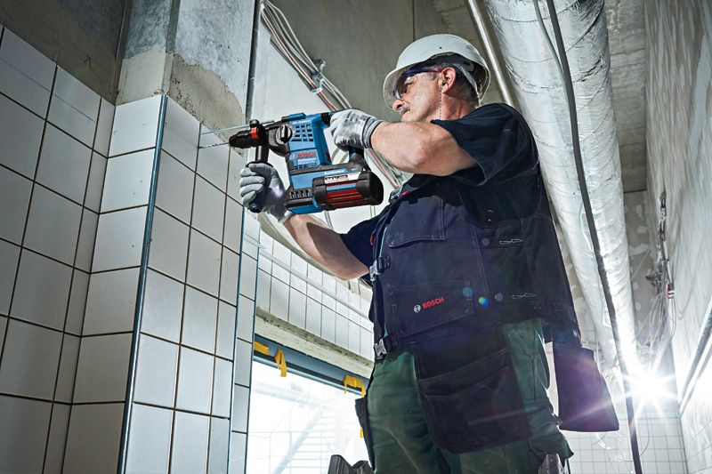 안전장비를 착용한 사람이 무선 회전 망치를 이용해 타일 벽에 구멍을 뚫고 있습니다.