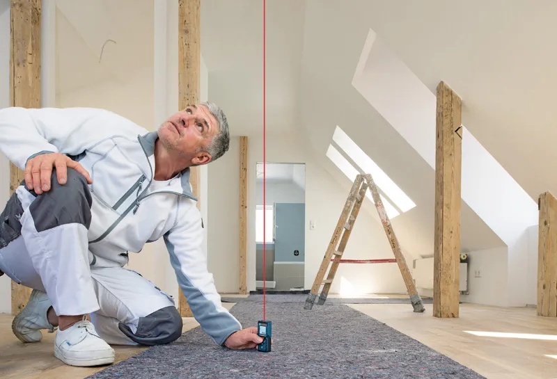 GLM 40-12 작업복을 입은 사람이 개조된 방에서 레이저 측정기를 사용하여 수직 거리를 측정하고 있습니다.