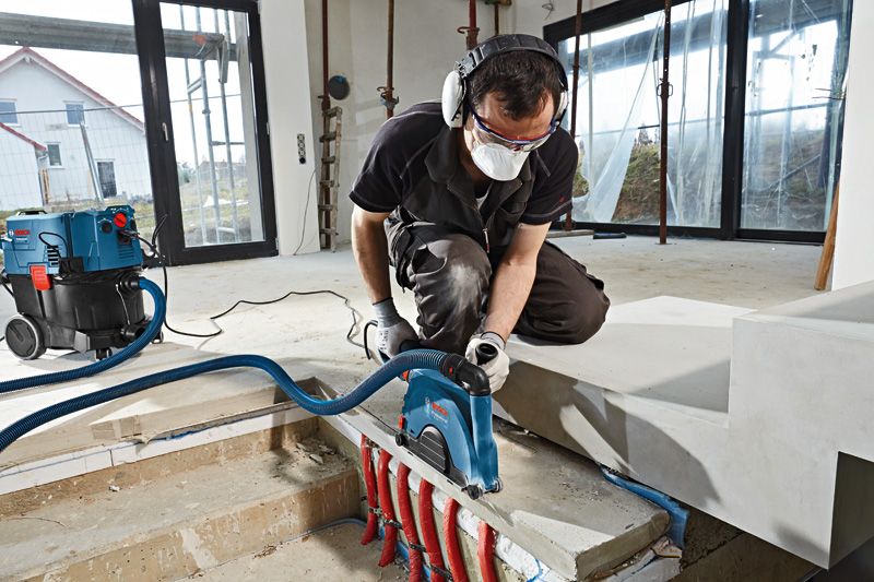 안전 장비를 착용한 사람이 벽면 절단기를 사용하여 노출된 배관 근처의 콘크리트를 절단하고 있습니다.