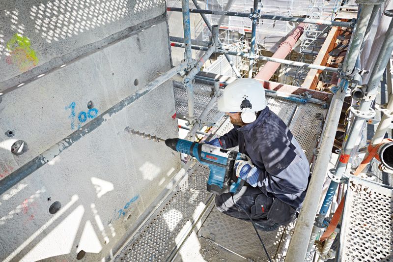 안전장비를 착용한 사람이 큰 회전 망치로 콘크리트를 뚫고 있습니다.