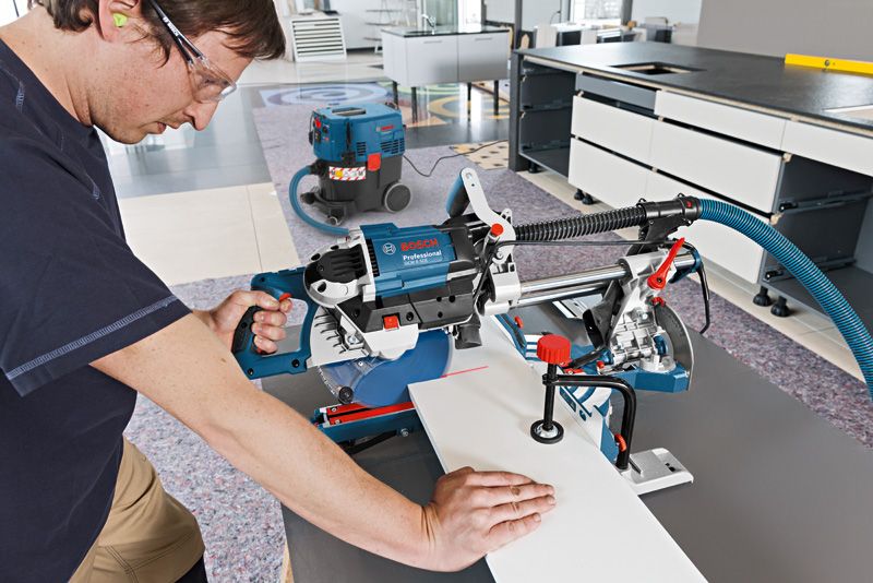 안전 장비를 착용한 사람이 작업장에서 슬라이딩 마이터 톱을 이용해 나무를 자르고 있습니다.