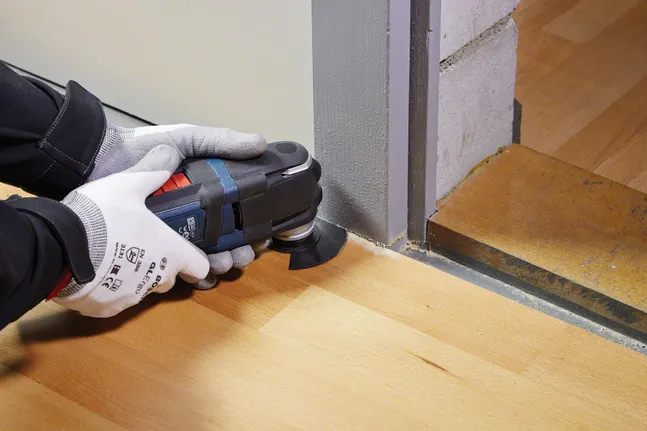 안전 장비를 착용한 사람이 문틀 근처에서 다기능 도구를 이용해 나무 바닥을 다듬고 있습니다.