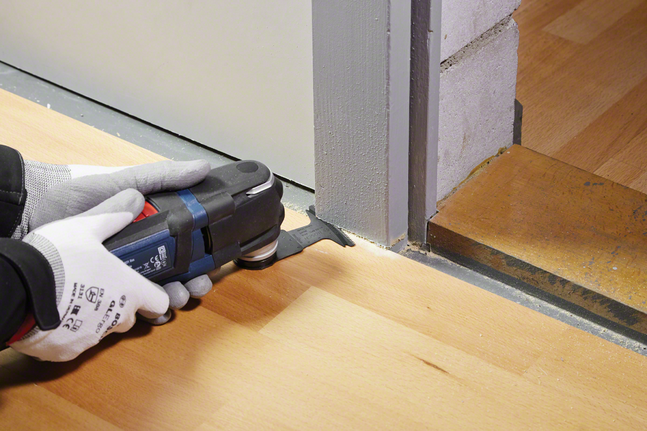 안전 장비를 착용한 사람이 전동 공구를 사용하여 문틀 옆의 나무 바닥을 다듬고 있습니다.