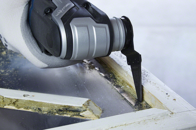 장갑을 낀 손으로 진동 도구를 이용해 오래된 창틀 퍼티를 자릅니다.