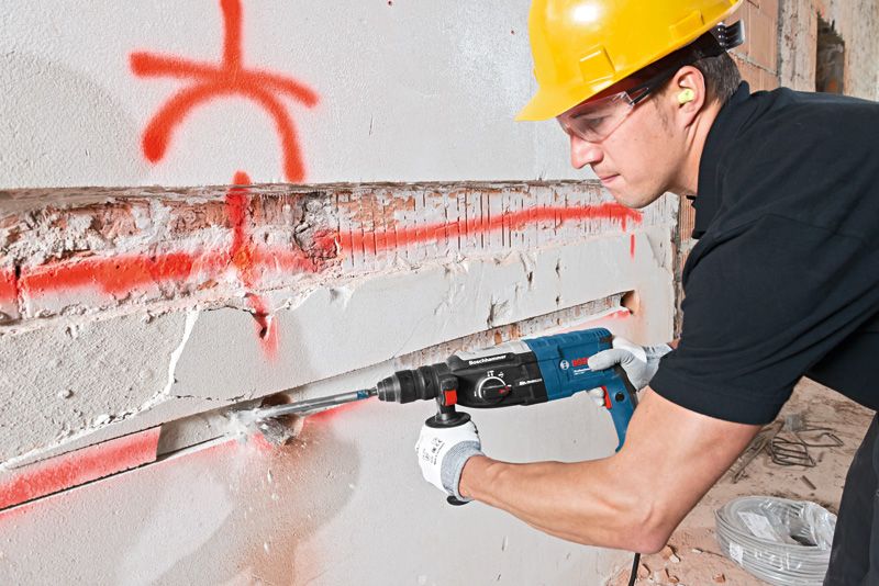 안전장비를 착용한 사람이 회전 망치를 사용해 콘크리트 벽에 홈을 파고 있습니다.