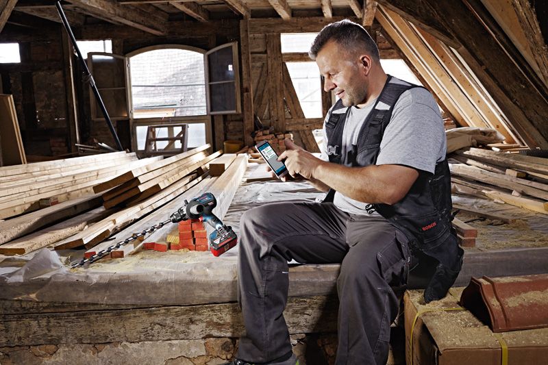 안전 장비를 착용한 사람이 목공 작업장에서 무선 드릴 옆에서 스마트폰을 사용하고 있습니다.