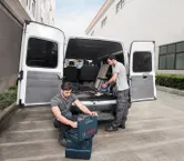 열린 밴 근처에 있는 두 명의 작업자가 도구를 정리하고 무선 진공 청소기를 사용하고 있습니다.