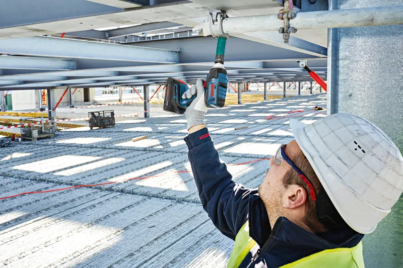 안전 장비를 착용한 사람이 무선 충격 렌치를 금속 빔에 사용하고 있습니다.