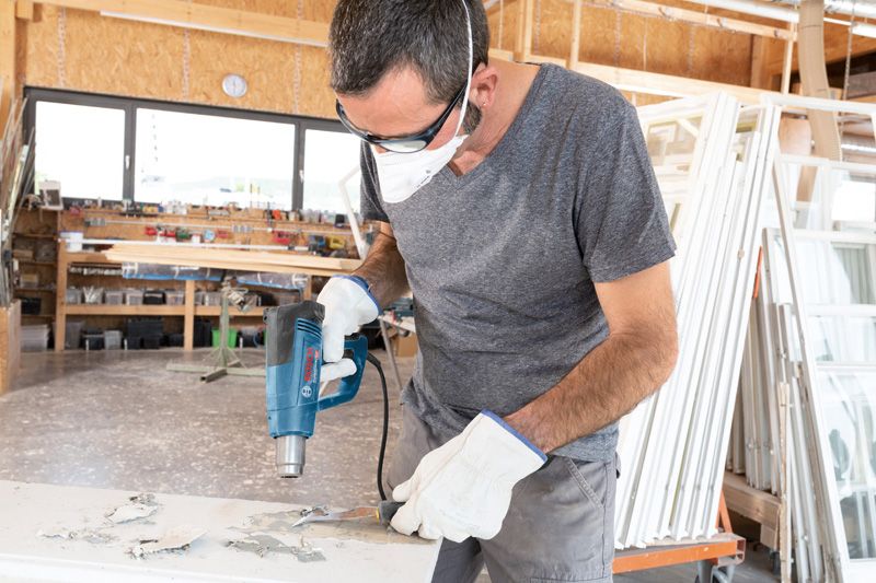 안전 장비를 착용한 사람이 열선총을 사용하여 표면의 페인트를 제거합니다.