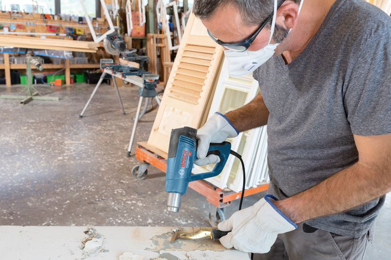 안전 장비를 착용한 사람이 열풍총을 사용하여 나무 표면의 페인트를 제거하고 있습니다.