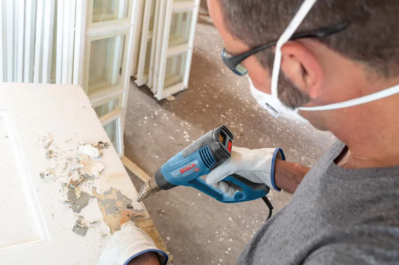 안전 장비를 착용한 사람이 열선총을 사용해 문의 페인트를 벗기고 있습니다.