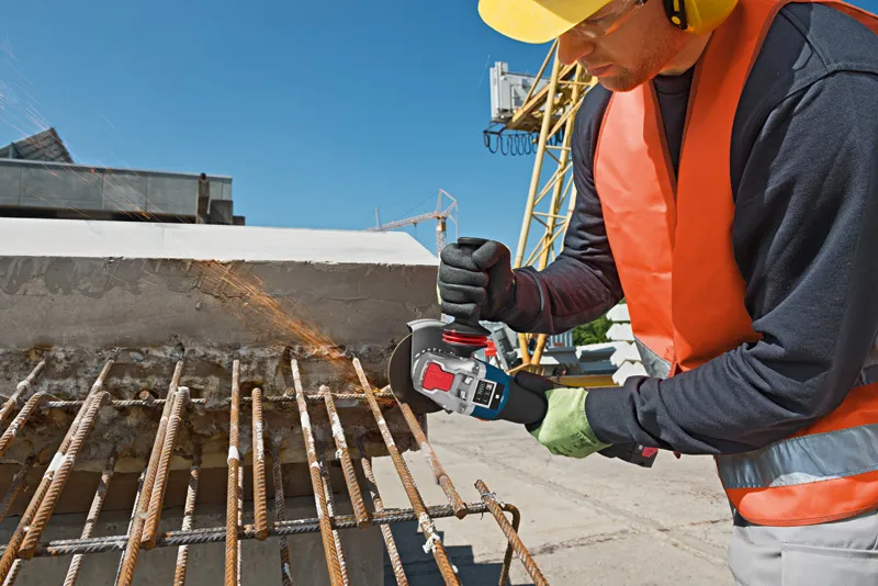 GWX 18V-10 SC 안전 장비를 착용한 사람이 건설 현장에서 각도 그라인더를 이용해 녹슨 철근을 자르고 있습니다.