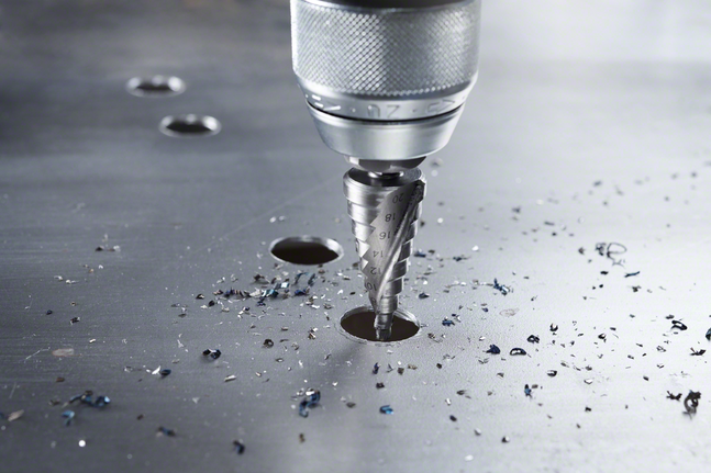 스텝 드릴 비트가 금속판에 구멍을 뚫고 그 사이로 드릴 ​​부스러기가 흩어지는 모습.