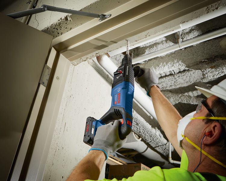 안전 장비를 착용한 사람이 무선 회전 망치를 이용해 콘크리트 천장에 구멍을 뚫고 있습니다.