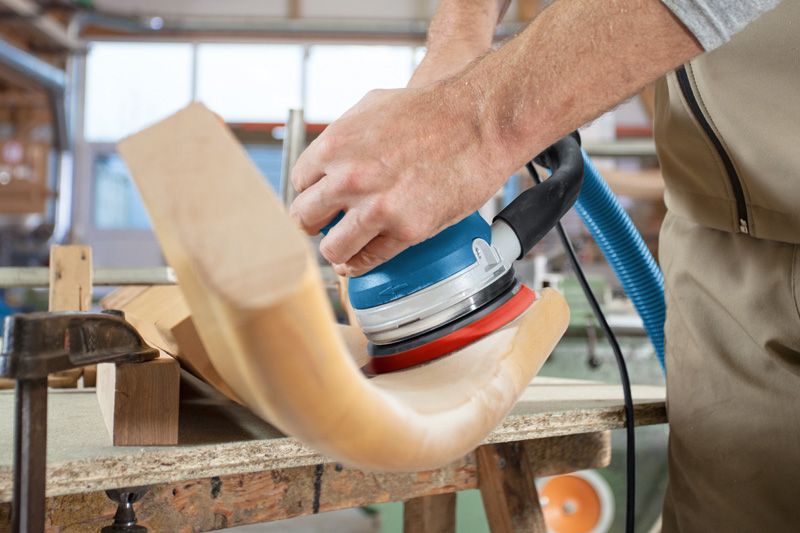 사람이 랜덤 오빗 샌더를 사용하여 곡선형 나무 의자 부분을 연마하고 있습니다.