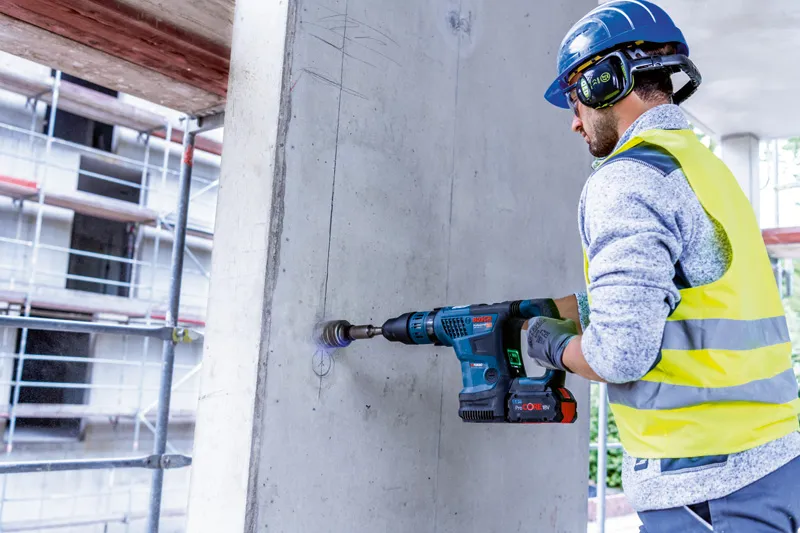 안전 장비를 착용한 사람이 무선 회전 망치로 콘크리트 벽에 구멍을 뚫고 있습니다.