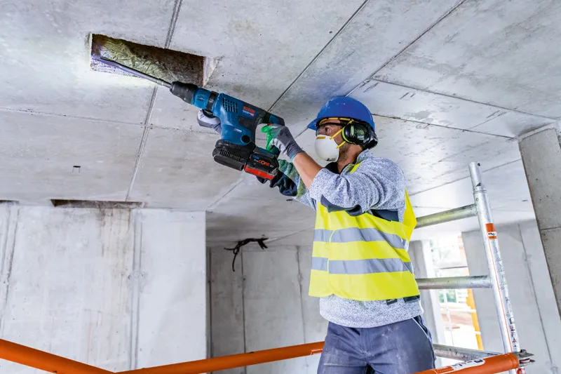 안전장비를 착용한 사람이 무선 회전 망치로 콘크리트 천장에 구멍을 뚫고 있습니다.