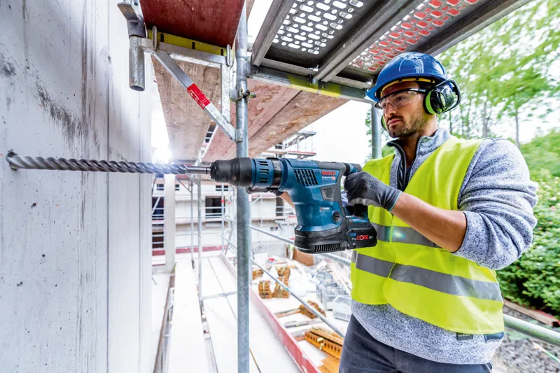 안전장비를 착용한 사람이 무선 회전 망치를 이용해 콘크리트에 구멍을 뚫고 있습니다.