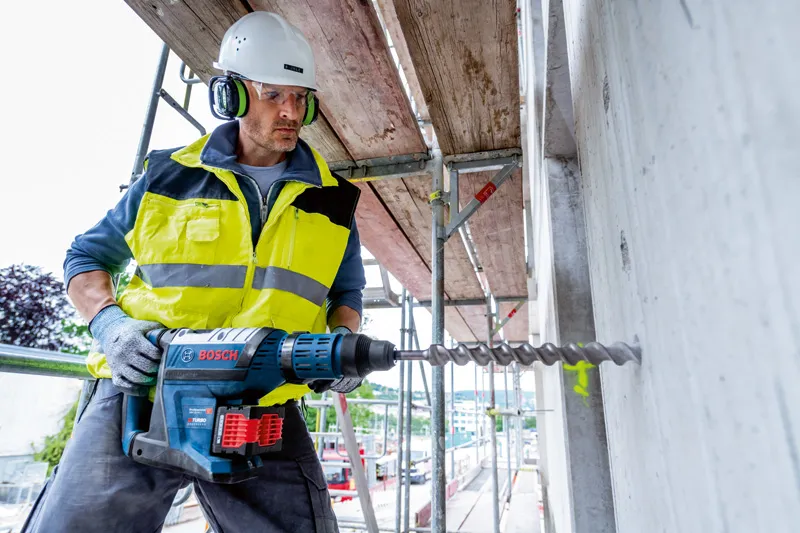 안전장비를 착용한 사람이 무선 회전 망치로 콘크리트를 뚫고 있습니다.