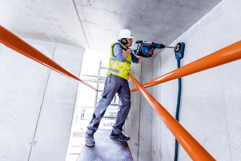 안전장비를 착용한 사람이 회전 망치로 콘크리트 천장에 구멍을 뚫고 있습니다.