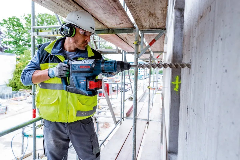 안전장비를 착용한 사람이 건설 현장에서 콘크리트 벽에 구멍을 뚫고 있다.