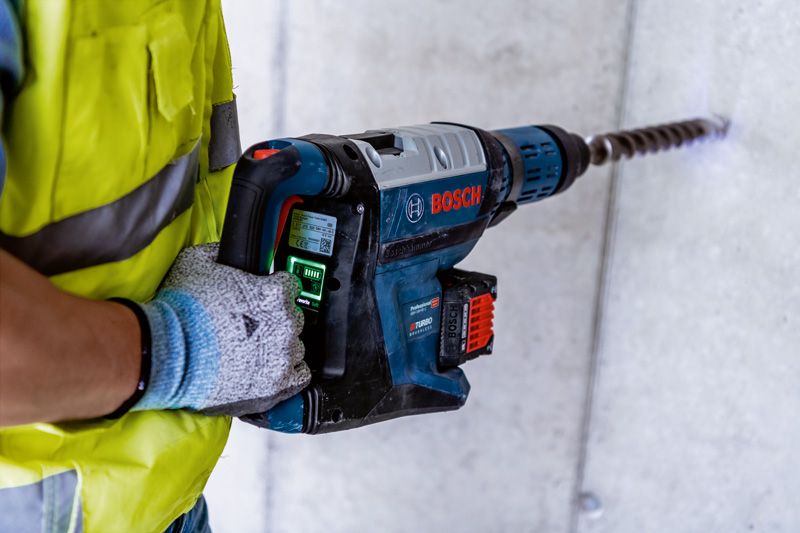 안전장비를 착용한 사람이 큰 회전 망치로 콘크리트를 뚫고 있습니다.