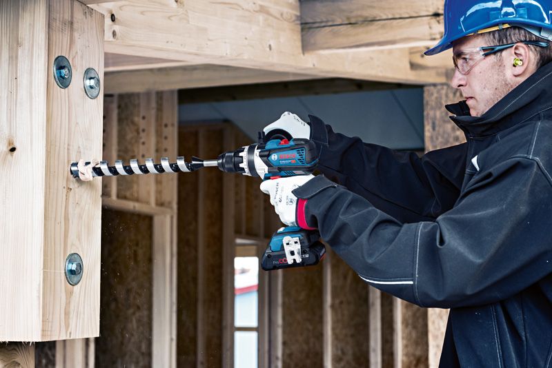 안전장비를 착용한 사람이 건설 현장에서 나무 들보에 구멍을 뚫고 있다.