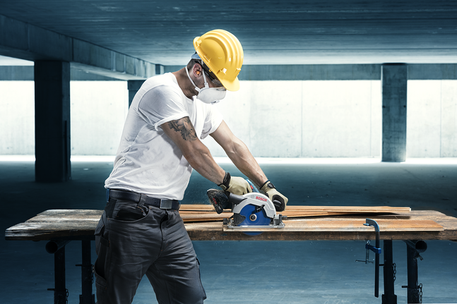 안전 장비를 착용한 사람이 작업대 위에서 원형 톱을 사용하여 나무를 자르고 있습니다.
