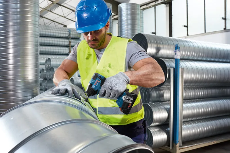 안전 장비를 착용한 사람이 무선 전동 공구를 사용하여 금속 배관을 고정하고 있습니다.
