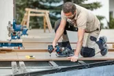 안전장비를 착용한 사람이 무선 드릴을 이용해 데크 보드를 고정하고 있습니다.
