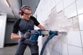 안전 장비를 착용한 사람이 무선 회전 망치를 사용하여 벽 타일을 제거하고 있습니다.