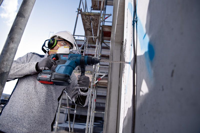 안전장비를 착용한 사람이 건설 현장에서 콘크리트 벽에 드릴을 박고 있다.