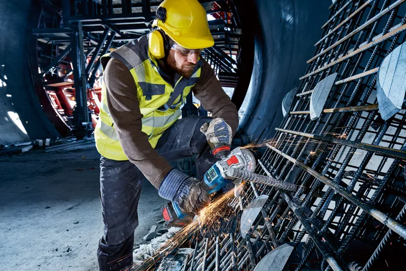 안전 장비를 착용한 사람이 무선 각도 그라인더를 사용하여 철근을 자릅니다.