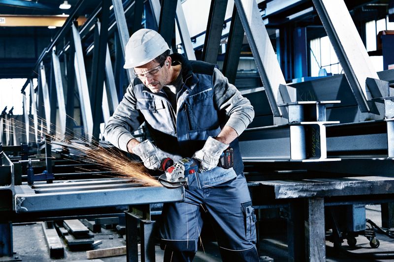 안전 장비를 착용한 사람이 무선 각도 그라인더로 금속 빔을 갈고 있습니다.