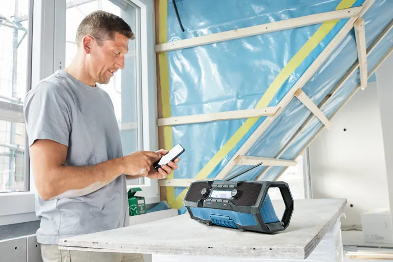 작업복을 입은 사람이 테이블 위의 작업 현장 라디오 옆에서 스마트폰을 조정하고 있습니다.