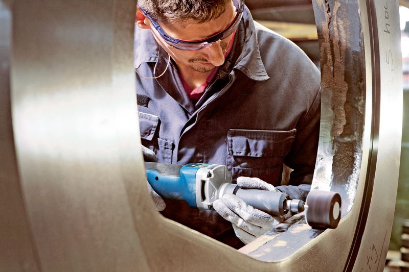 안전 장비를 착용한 사람이 무선 스트레이트 그라인더로 큰 금속 링을 닦고 있습니다.