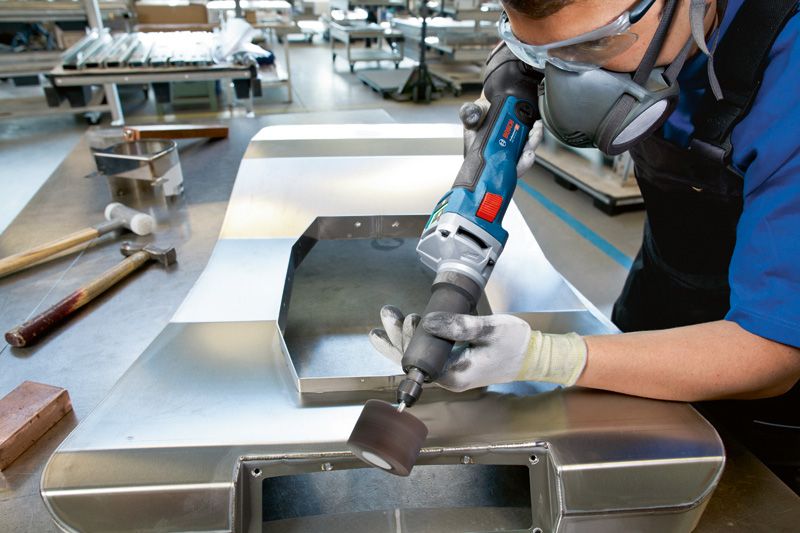 안전 장비를 착용한 사람이 무선 스트레이트 그라인더로 금속판을 갈고 있습니다.