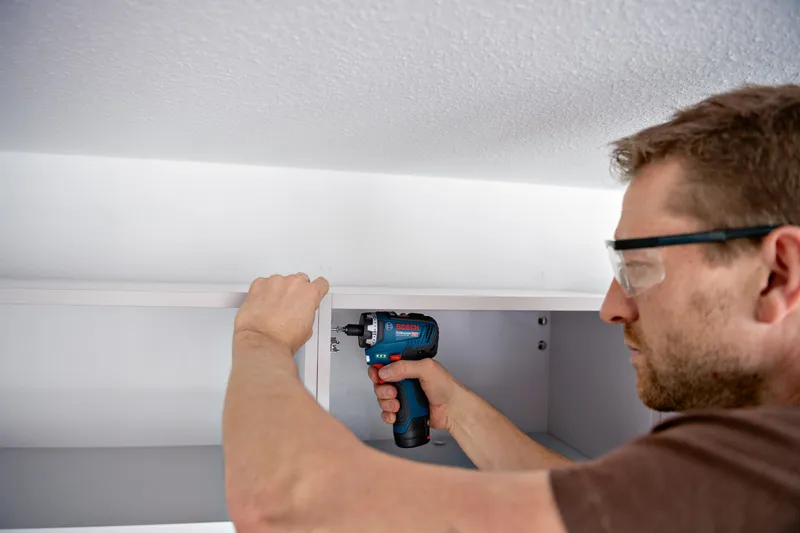 안전장비를 착용한 사람이 무선 드라이버를 사용하여 캐비닛을 조립하고 있습니다.