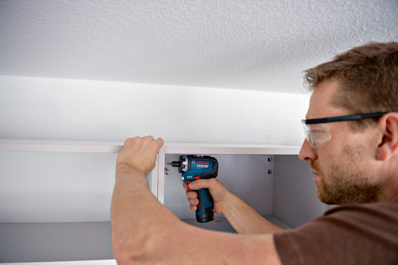 안전장비를 착용한 사람이 무선 드라이버를 사용하여 캐비닛을 조립하고 있습니다.