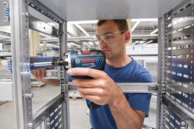 안전 장비를 착용한 사람이 금속 프레임에 무선 드라이버를 사용하고 있습니다.