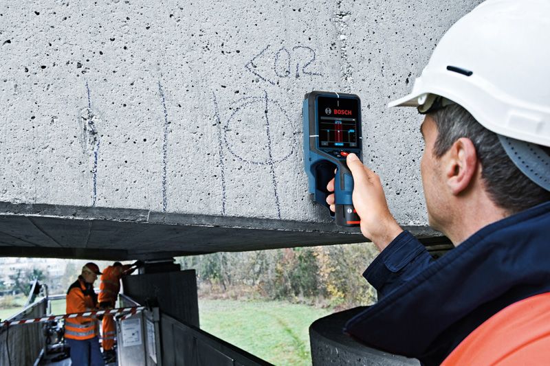 안전 장비를 착용한 사람이 콘크리트 기둥에 탐지기를 사용하고 있습니다.