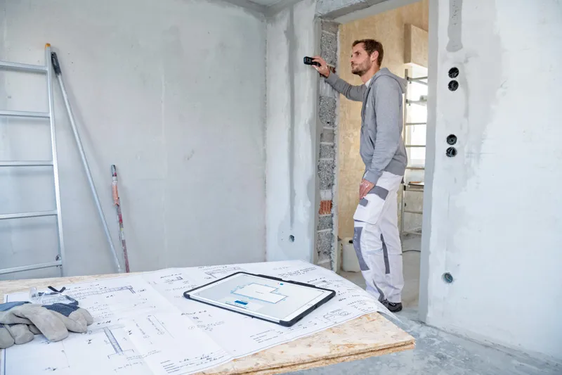 한 사람이 건설 현장에서 레이저 측정기를 사용하여 벽 거리를 확인하고 있습니다.