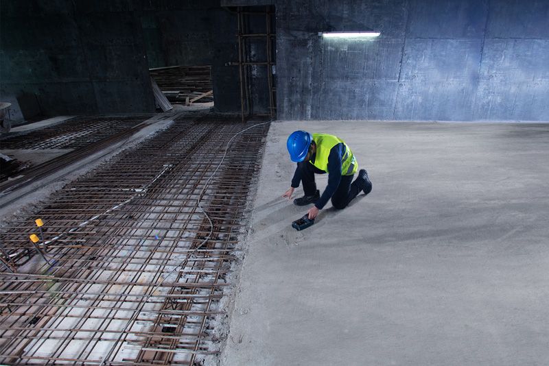 안전 장비를 착용한 사람이 노출된 철근 근처에서 탐지기로 콘크리트를 검사합니다.