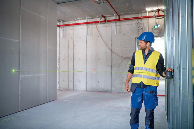 안전 장비를 착용한 사람이 레이저 레벨링 도구를 사용하여 벽 프레임을 정렬하고 있습니다.