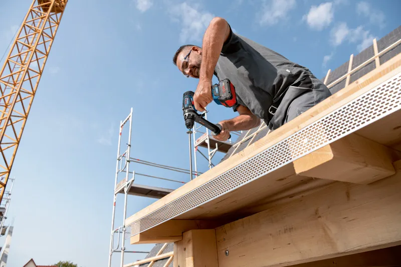 GSB 18V-150 C 안전장비를 착용한 사람이 건설 중인 건물의 나무 들보에 구멍을 뚫고 있습니다.