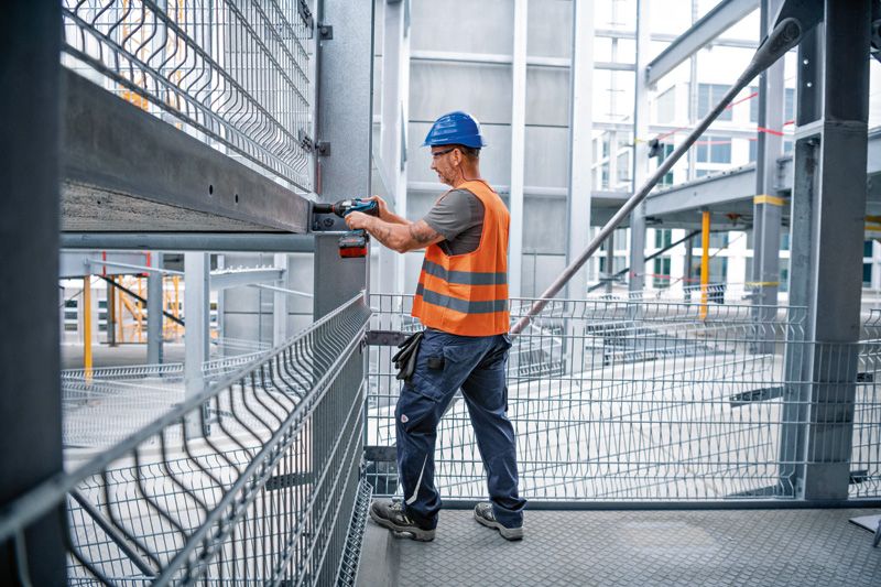 GDS 18V-1000 C 안전 장비를 착용한 사람이 무선 충격 렌치를 사용하여 강철 보를 작업하고 있습니다.